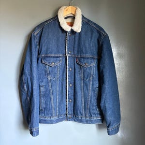 May include: A blue denim jacket with a cream-colored sherpa collar and lining. The jacket has two chest pockets, two side pockets, and a button-up front. A red tag is visible on the chest pocket.