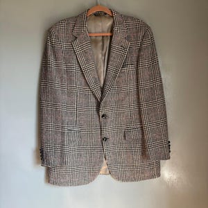 May include: A brown, black, and white checked blazer with a two-button fastening and notched lapels. The blazer is suspended on a wooden hanger. It features two front pockets and buttoned cuffs. The background is grey.