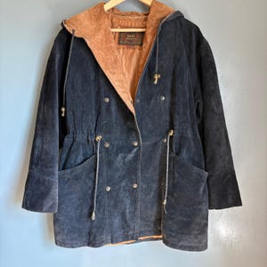 Könnte beinhalten: Dunkelmarineblaue Wildlederjacke mit brauner Wildleder-Kapuze und -Futter. Die Jacke hat einen doppelreihigen Verschluss mit goldfarbenen Knöpfen, eine Taille mit Kordelzug und zwei Vordertaschen. Die Jacke hängt an einem Holzbügel.