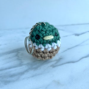 May include: A small crocheted duck keyring made with green, white and brown yarn. The duck has black eyes and a yellow beak.
