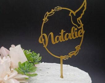 Pode incluir: Topper de bolo dourado com um design circular, uma pomba e o nome "Natalie" em itálico. O topper está em um bolo branco com flores rosa e verdes.