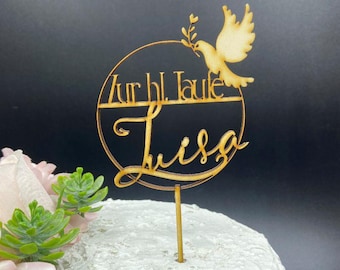 Pode incluir: Topper de bolo de madeira com as palavras "Zur hl. Taufe Luisa" num design circular, apresentando uma pomba carregando um ramo de oliveira. O topper está em um bolo branco com cobertura, com vegetação e flores rosas em primeiro plano.