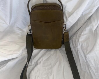 May include: Olive green crossbody bag with a top handle and a long, adjustable strap. The bag has a zippered main compartment and a small front pocket. The hardware is silver-toned. The bag is made of a textured material.