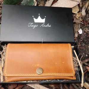 May include: A brown leather wallet with a gold button closure. The wallet is in a black box with a white crown and the text "Tiago Andre" on the lid.