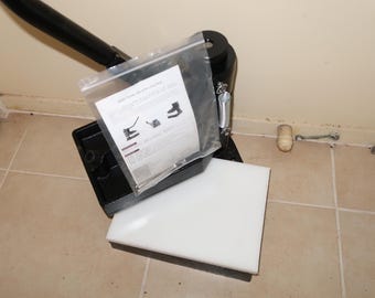 May include: Black metal paper trimmer with a white cutting mat and instructions. The trimmer has a spring-loaded blade and a safety guard.