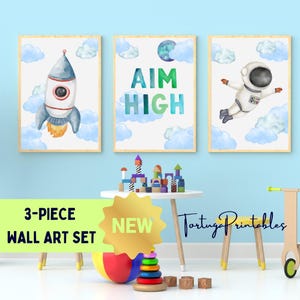 Puede incluir: Un conjunto de arte mural de tres piezas con ilustraciones de acuarela. Una impresión muestra un cohete, otra dice "AIM HIGH", y la tercera representa a un astronauta en el espacio. Las impresiones están enmarcadas en madera clara.