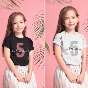 May include: Two girls wearing white and black t-shirts with the number 5 and the name "Sophia" on them. The girl on the left is wearing a black t-shirt with a pink number 5 and the girl on the right is wearing a white t-shirt with a silver glitter number 5.
