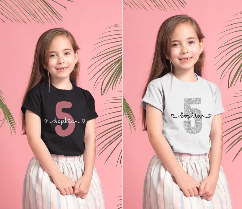 May include: Two girls wearing white and black t-shirts with the number 5 and the name "Sophia" on them. The girl on the left is wearing a black t-shirt with a pink number 5 and the girl on the right is wearing a white t-shirt with a silver glitter number 5.