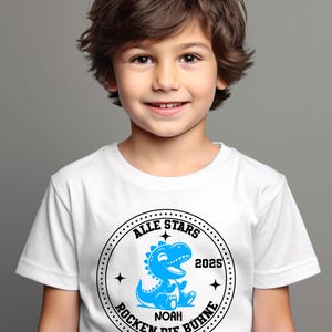 May include: White t-shirt with a black and blue circular graphic. The graphic features a blue dinosaur with the text "Alle Stars 2025 Rocken Die B&uuml;hne" and "Noah" in the center.