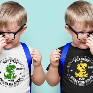 May include: Two children wearing black framed glasses and blue backpacks are wearing white and black t-shirts with a green and yellow dinosaur graphic. The shirts say "Alle Stars" and "Rocken die Buhne" with the year 2025.
