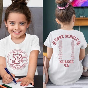 May include: White t-shirt with a red and white graphic of a koala bear with the text "ALLE STARS 2025 EMILIA ROCKEN DIE BUHNE". The back of the shirt has the text "EURE SCHULE" with a list of names and "KLASSE 4A".