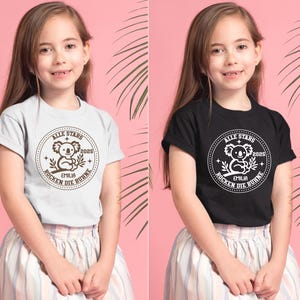 May include: Two children wearing t-shirts with a brown koala graphic and the text "ALLE STARS 2025 EMILIA ROCKEN DIE B&Uuml;HNE" in a circle. The girl on the left is wearing a light gray t-shirt and the girl on the right is wearing a black t-shirt.