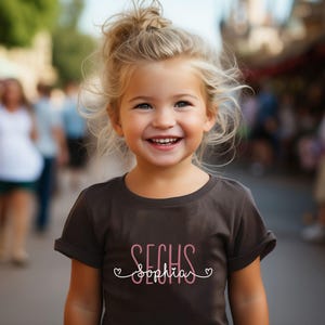 Peut inclure: Une jeune fille portant un t-shirt marron avec le texte "SECHS Sophia" en lettres roses. La fille sourit et a les cheveux blonds.