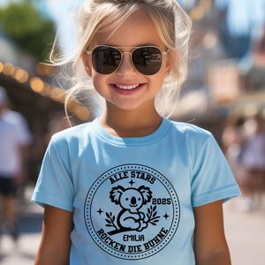 May include: A light blue t-shirt with a black and white graphic of a koala bear inside a circle. The circle has the text "ALLE STARS" at the top, "ROCKEN DIE B&Uuml;HNE" at the bottom, and "2025" to the right of the koala. The text "EMILIA" is below the koala.