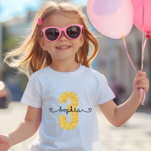 May include: A white t-shirt with a gold glitter number 3 and the name "Sophia" written in black cursive. The shirt is being worn by a young girl with pink sunglasses and holding pink balloons.