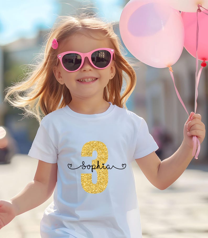 May include: A white t-shirt with a gold glitter number 3 and the name "Sophia" written in black cursive. The shirt is being worn by a young girl with pink sunglasses and holding pink balloons.