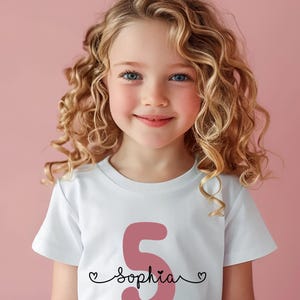 Peut inclure: Un t-shirt blanc avec un chiffre 5 rose et le nom "sophia" &eacute;crit en cursive noire avec un c&oelig;ur de chaque c&ocirc;t&eacute; du nom.