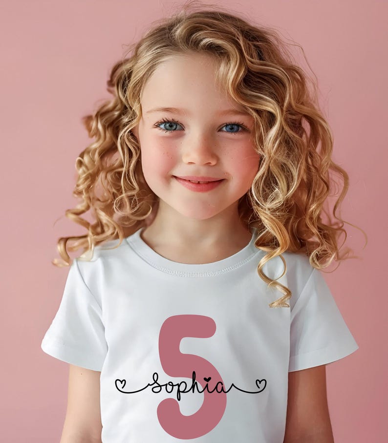Peut inclure: Un t-shirt blanc avec un chiffre 5 rose et le nom "sophia" &eacute;crit en cursive noire avec un c&oelig;ur de chaque c&ocirc;t&eacute; du nom.