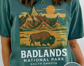 May include: Blue-spruce colored t-shirt featuring a graphic of the Badlands National Park in South Dakota. The design includes a bison, mountains, and the text "BADLANDS NATIONAL PARK SOUTH DAKOTA".
