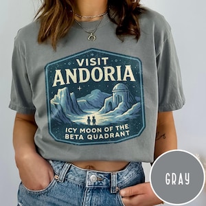 May include: Gray t-shirt with a blue and white graphic that reads "VISIT ANDORIA, ICY MOON OF THE BETA QUADRANT." The graphic features a landscape with mountains, a building, and two figures. The shirt is a solid gray color.