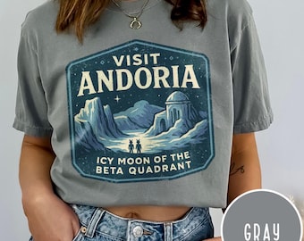 May include: Gray t-shirt with a blue and white graphic that reads "VISIT ANDORIA, ICY MOON OF THE BETA QUADRANT." The graphic features a landscape with mountains, a building, and two figures. The shirt is a solid gray color.