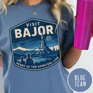 May include: Blue t-shirt with a graphic that says "Visit Bajor" with a landscape design. The design includes a tower, mountains, and the text "Heart of the Spiritual Life". A pink tumbler is held in the right hand.