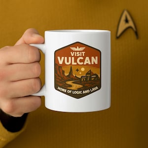 May include: White ceramic mug featuring a retro-style travel design with the text "Visit Vulcan" and "Home of Logic and Lava." The design includes a landscape with mountains, a river, and buildings in shades of brown, orange, and yellow.