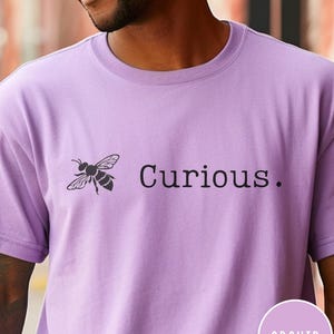 May include: A purple t-shirt with a black bee graphic and the text "Curious." printed on the front. The shirt is a unisex style.