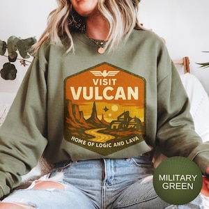 May include: Military green sweatshirt with a graphic that says "Visit Vulcan" in orange and white. The graphic depicts a desert landscape with a winding road and buildings. The text "Home of Logic and Lava" is also included.