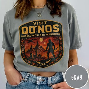 May include: Gray t-shirt with a graphic featuring the text "VISIT QO'NOS RUGGED WORLD OF WARRIORS." The design includes a warrior silhouette, a mountain landscape, and a stylized border in shades of orange and black.