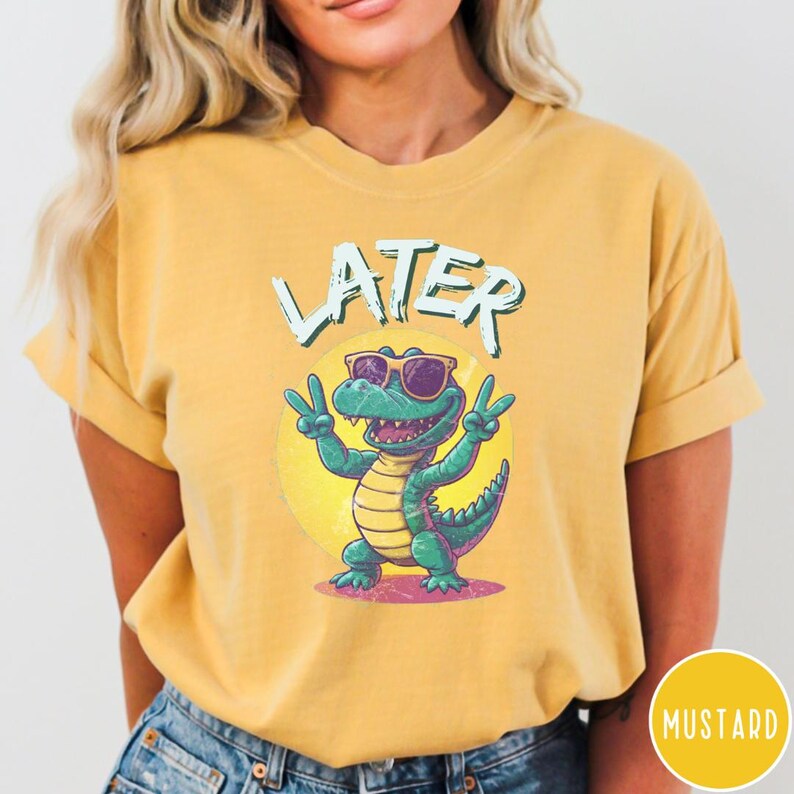 May include: A yellow t-shirt with a cartoon alligator wearing sunglasses and the word "LATER" printed on it. The alligator is wearing sunglasses and has a peace sign with each hand.