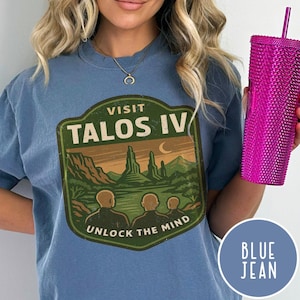 May include: A blue jean colored t-shirt with a graphic that says "Visit Talos IV" with a landscape design. The shirt is being held by a person holding a pink studded tumbler. The text "Unlock the Mind" is below the landscape graphic.