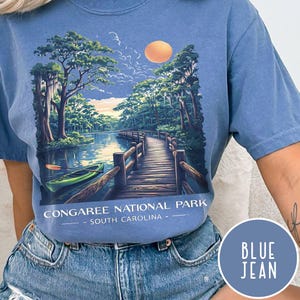May include: Blue jean-colored t-shirt featuring a scenic illustration of Congaree National Park in South Carolina. The design includes a wooden bridge over a river, a kayak, trees, and a sunset. Text on the shirt reads "CONGAREE NATIONAL PARK - SOUTH CAROLINA".