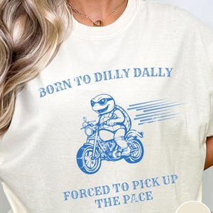 May include: A white t-shirt with blue text that reads "Born to dilly dally, forced to pick up the pace". A cartoon turtle wearing sunglasses is riding a motorcycle. The word "Ivory" is in the bottom right corner.