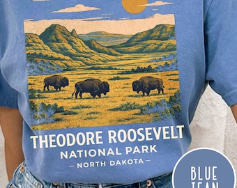 May include: Blue t-shirt featuring a scenic illustration of Theodore Roosevelt National Park. The design includes bison grazing in a field, mountains, and the text "THEODORE ROOSEVELT NATIONAL PARK - NORTH DAKOTA -."