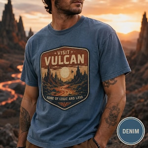 May include: A blue short-sleeve t-shirt featuring a graphic patch with the text VISIT VULCAN HOME OF LOGIC AND LAVA. The design depicts a landscape with jagged mountains, a glowing sun, and a winding path in shades of orange, brown, and cream.