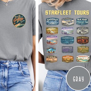 May include: A grey t-shirt featuring a small circular graphic on the front chest that says Visit The Galaxy. The back displays the text STARFLEET TOURS above fifteen rectangular travel-style patches depicting various fictional planetary locations.