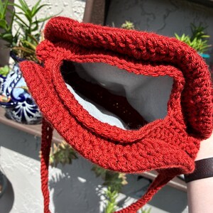 May include: A handmade, red crochet bag with a textured stitch pattern. The bag has a rectangular opening and a long, braided strap. The interior of the bag is lined with a light-colored fabric. The bag is shown outdoors.