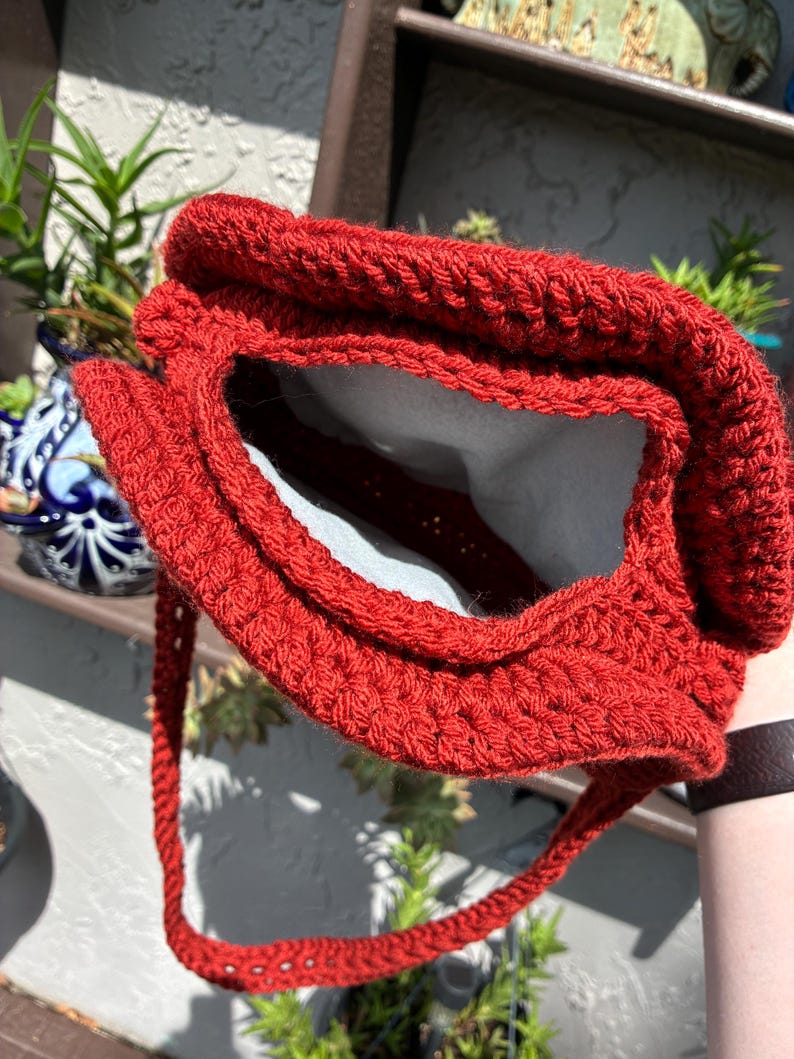 May include: A handmade, red crochet bag with a textured stitch pattern. The bag has a rectangular opening and a long, braided strap. The interior of the bag is lined with a light-colored fabric. The bag is shown outdoors.