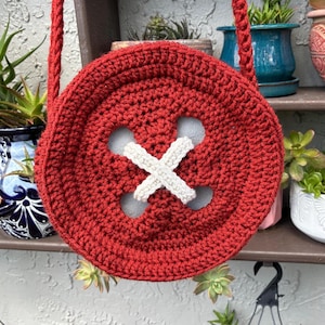 May include: A handmade, red crocheted button-shaped bag with a white cross-stitch detail. The bag has a matching red strap for over-the-shoulder or crossbody wear. The bag is round and has a unique, whimsical design.