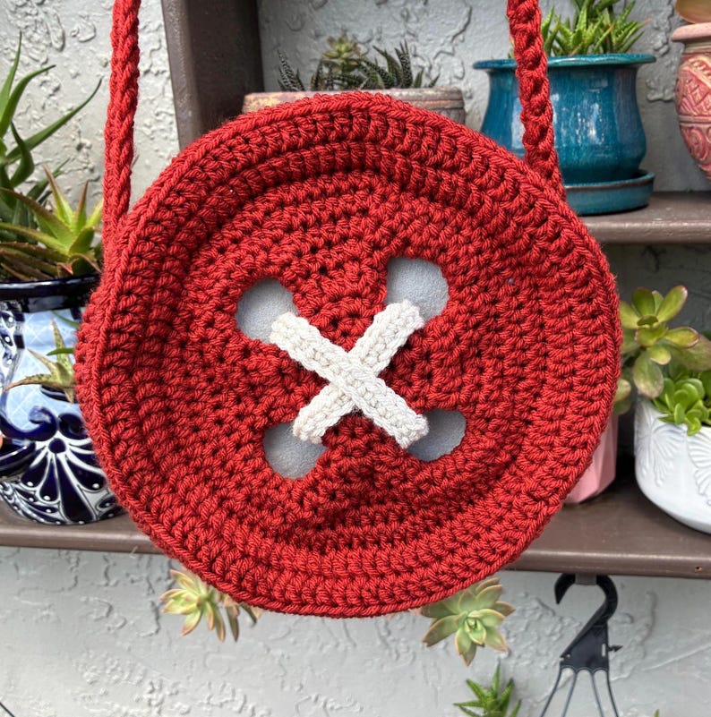May include: A handmade, red crocheted button-shaped bag with a white cross-stitch detail. The bag has a matching red strap for over-the-shoulder or crossbody wear. The bag is round and has a unique, whimsical design.