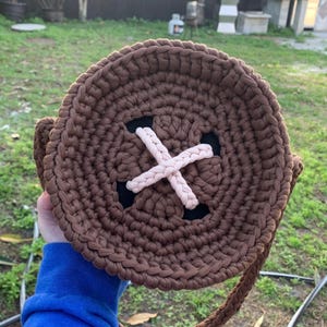 Puede incluir: Un bolso redondo de ganchillo marrón con un diseño de cruz rosa claro en el centro. El bolso tiene una correa trenzada y detalles en negro. El bolso es hecho a mano y tiene una apariencia texturizada única.