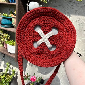 May include: A red crocheted button-shaped purse with a white 'X' design in the center. The purse has a long, braided strap and is held up against a background of plants and a wall.