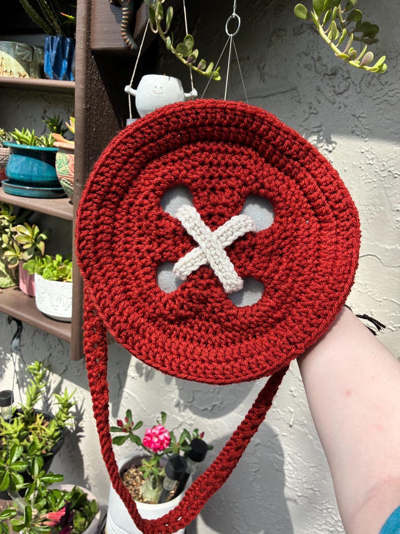 May include: A red crocheted button-shaped purse with a white 'X' design in the center. The purse has a long, braided strap and is held up against a background of plants and a wall.
