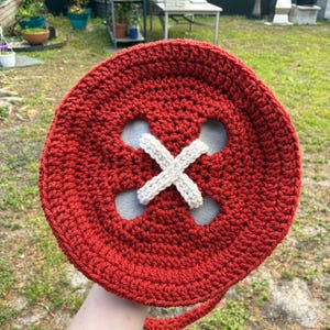 May include: A large, round, red crocheted bag resembling a button. The bag features four holes with a cream-colored, crocheted 'X' design in the center. A matching red strap extends from the side. The bag is handmade.