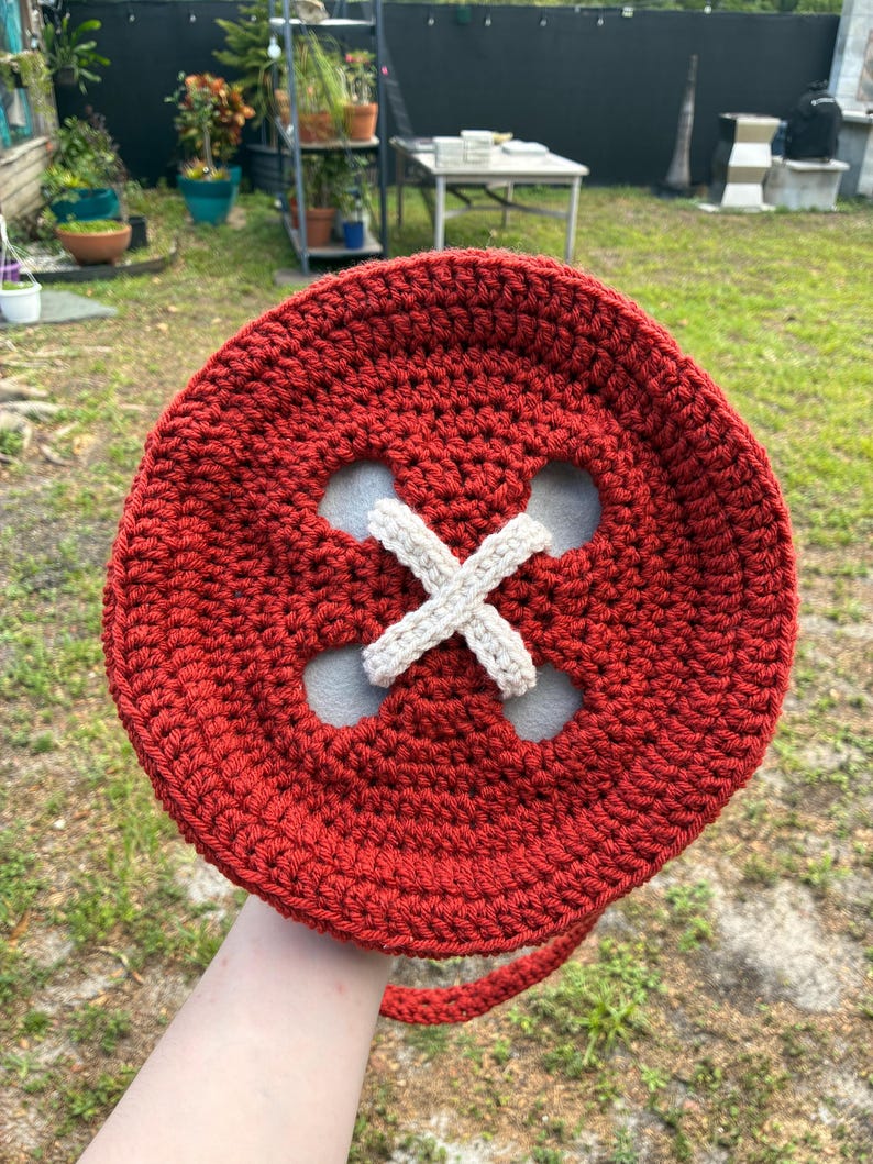 May include: A large, round, red crocheted bag resembling a button. The bag features four holes with a cream-colored, crocheted 'X' design in the center. A matching red strap extends from the side. The bag is handmade.