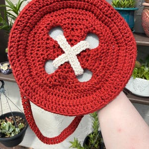 May include: A handmade, red crocheted button-shaped bag with a cream-colored cross design in the center. The bag has a matching red strap and is held up against a background of plants.