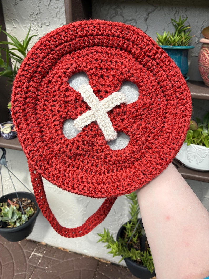 May include: A handmade, red crocheted button-shaped bag with a cream-colored cross design in the center. The bag has a matching red strap and is held up against a background of plants.