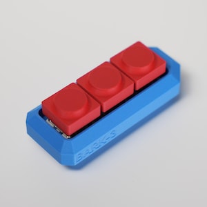 Peut inclure: Un objet rectangulaire bleu et rouge avec trois boutons carrés rouges avec des dessus circulaires. La base est bleue et les boutons sont rouges. Le texte "BARK-S" est visible sur le côté.