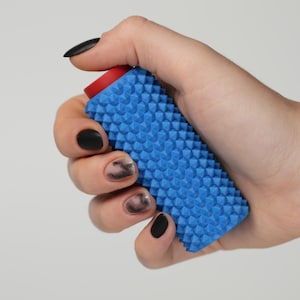 Peut inclure: Un rouleau de massage bleu avec une surface texturée et un capuchon rouge est tenu dans une main avec du vernis à ongles noir. La surface du rouleau est recouverte de petites bosses en forme de cône. Le fond est gris clair uni.
